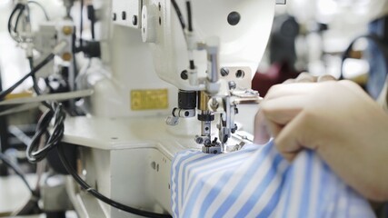 Sewing on machine, hands and blue and white striped cloth close up. Sewing process, female hands on white machine, blue shirt on table with white sewing machine