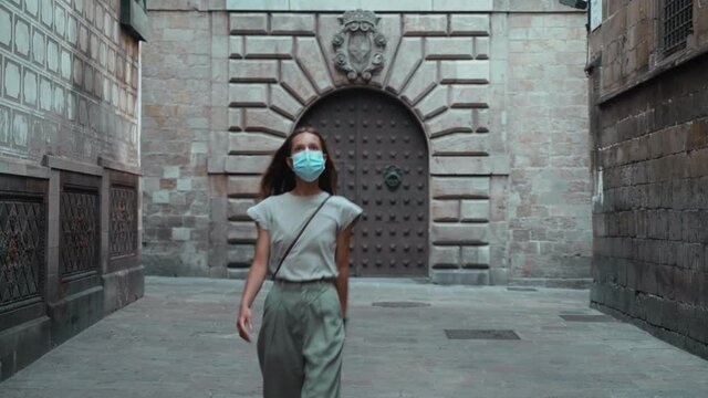 Beautiful long-haired brunette in protective mask walking around the beautiful city. Young attractive woman walk the stone streets of a medieval town. COVID-19 precaution. Stay safe. Pretty tourist.