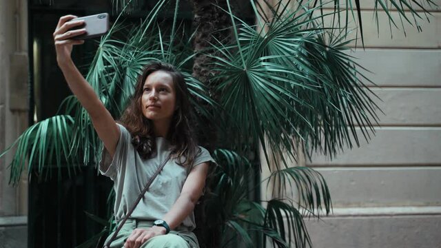 Beautiful long-haired brunette taking selfie while sitting on a street pot with a palm tree. Young attractive woman with the cellphone on the street. Pretty tourist taking pictures on a mobile phone.