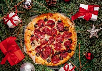 Christmas pepperoni pizza with salami sausages, mozzarella, olives, Provencal herbs, branded tomato sauce, with Christmas decorations, gifts, spruce, toys