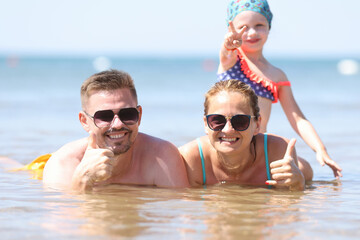 Fototapeta premium Family vacation with children at sea. Man and woman in sunglasses lie in water and show thumbs up. Girl sit on top of her mother and show two fingers.