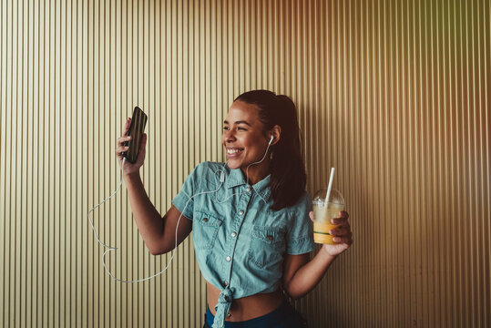 Cheerful woman holding smart phone and drink while enjoying music through headphones against wall