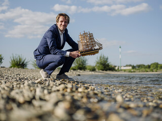 Smiling businessman holding toy boat at riverbank against sky during sunny day