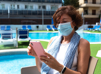 Woman with surgical mask consulting her mobile, vacation setting with swimming pool. concept of new...