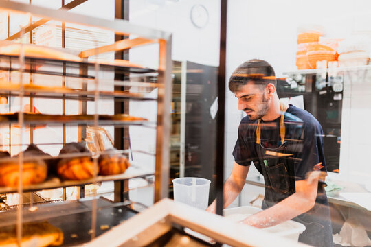 Young baker working at bakery seen through glass window