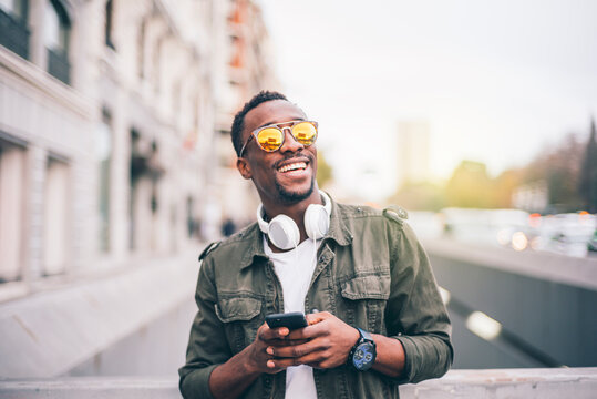 Cheerful man wearing sunglasses using smart phone while standing in city