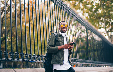 Smiling young man wearing sunglasses using mobile phone while standing against fence in city