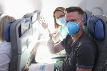 Man with protective mask sit on plane and show his thumbs up. Woman sit next to her husband and...