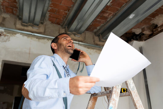 Laughing architect with plan on the phone in a house under construction