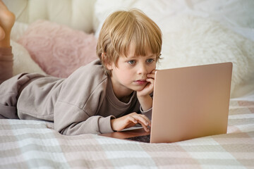 Cute child boy doing homework lying on bed at home. Studying online , distance learning, self education       