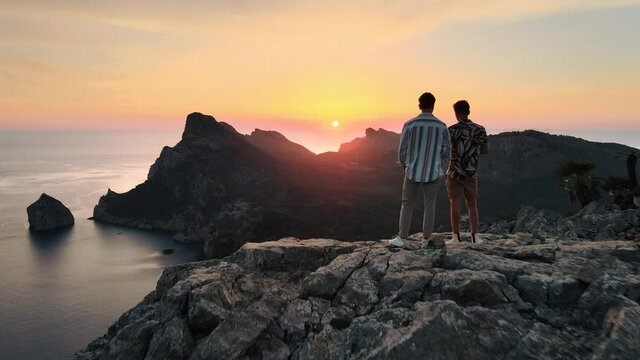 Aerial dolly out shot revealing two friends standing together on top of a high cliff in Mallorca and looking the sunrise
