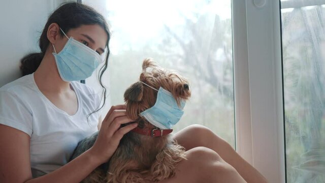 coronavirus pandemic concept. little teenage girl sitting in a medical mask at the window puts on a mask to the dog. self-isolation concept virus covid 19 infection doomsday. sick child kid indoors