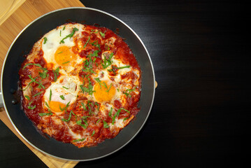 Shakshuka - poached eggs in tomato sauce, onion, pepper and spices in iron pan on dark wooden table with copy space. Famous traditional Arabic and Israeli breakfast - chakchouka. Top view.