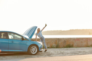 Full length shot of young cheerful woman smiling aside and showing peace sign while trying to repair her broken car, standing alone on the road side