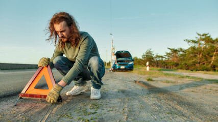 Man driver putting red warning triangle or emergency stop sign behind his broken car on the side of the road, his girlfriend waiting in the background