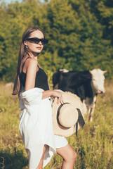 
A slender woman of model appearance in stylish glasses holds a hat in her hands and is in nature near a cow