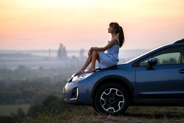 Yong happy female driver resting on hood of her car enjoying sunset view of summer nature. Travel destinations and recreation concept.