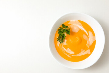 A bowl of pumpkin or carrot vegetarian cream soup is garnished with fresh parsley and cream on a white textured table. flat lay, copy space