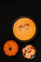 A bowl of pumpkin or carrot cream soup is decorated with shrimp and cream on a black background. flat lay, copy space.