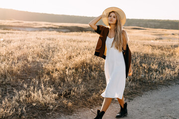Young blonde woman wearing a vintage dress and a straw hat, walking in countryside.
