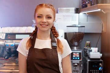 Fototapeta na wymiar Red haired barista posing in cafe shop, wearing white t shirt and brown apron, standing with coffee machine on background, looking smiling at camera.