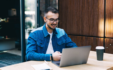 Happy young man in eyewear browsing netbook in summer area of urban cafeteria