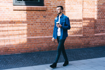 Full body of stylish young man walking near brick building with coffee cup