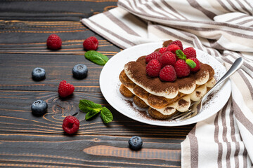 portion of Classic tiramisu dessert with raspberries and blueberries on wooden background