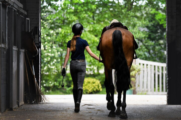 The back of the rider, walking towards the exit with her horse by her side with saddle on, ready to ride