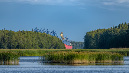Obraz premium Dry cargo vessel with red-orange hull and gantry cranes of coal handling terminal in Vysotsk cargo port viewed from fairway through strait between islands in Vyborg Bay.