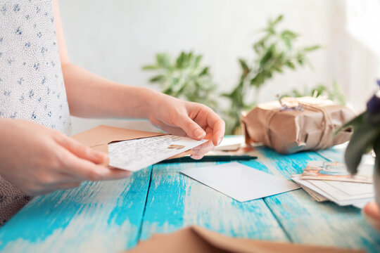 A woman holding a postcard with a craft envelope. Hands close-up. Blue wooden table with a parcel in the background. Concept of postcrossing and communication