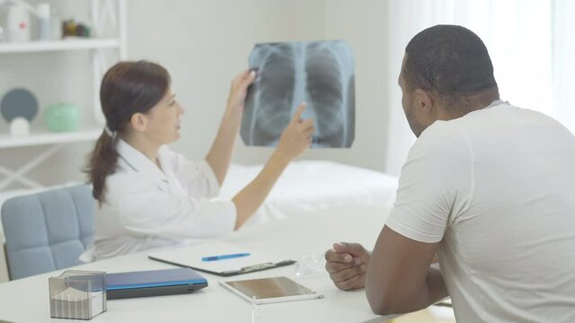 Professional Caucasian doctor explaining complications on lungs x-ray to African American patient. Confident woman talking with ill man in hospital. Consequence of chronic disease or smoking.