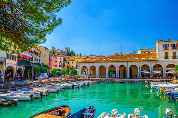 Old harbour Porto Vecchio with motor boats on turquoise water, green trees and traditional buildings in historical centre of Desenzano del Garda town, blue sky background, Lombardy, Northern Italy