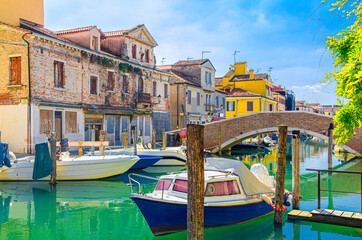 Fototapeta na wymiar Chioggia cityscape with narrow water canal with moored multicolored boats and yachts between old colorful buildings and brick bridge, blue sky background in summer day, Veneto Region, Northern Italy