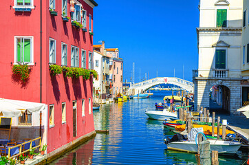 Fototapeta na wymiar Stone bridge Ponte di Vigo across Vena water canal with colorful boats and old buildings in historical centre of Chioggia town, blue sky background in summer day, Veneto Region, Northern Italy