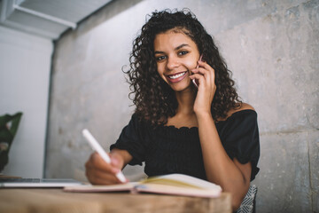 Portrait of prosperous female student sitting in coworking and phoning to friend for discussing education plan, cheerful African American woman with cute smile on face enjoying mobile conversation