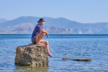 Senior asian man in face mask sits alone on a stone in a sea bay