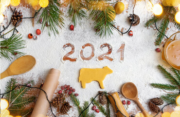 Happy New Year 2021 written on flour and Christmas tree branches. Ox-shaped cookies, cinnamon, spices, red berries, wooden spoons, parchment on a wooden background. Christmas card, new year card