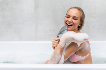 Young woman sing a song holding shower handle in hands in foam in bathroom.