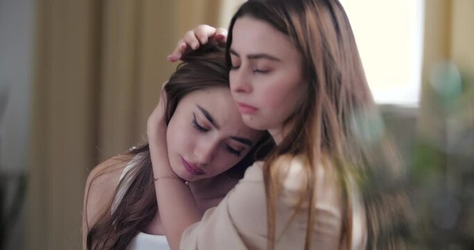 Close up young woman consoles sad unhappy friend, hugs sister by shoulders, presses support. Two teenage girls sitting at home on sofa discussing problems, stress and friendly concern care concept