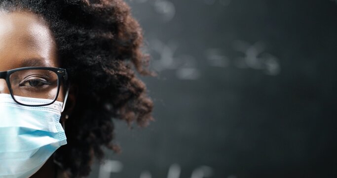 Close up of half face of young African American female teacher in glasses and medical mask looking at camera. Blackboard with math and physics formulas on background. School during pandemia. Portrait.