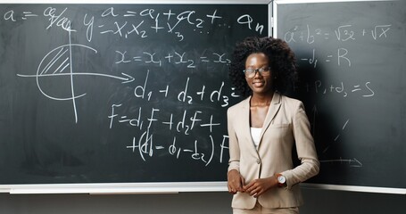 Portrait of young African American female teacher in glasses standing at board, writing with chalk math or physics laws and formulas and smiling to camera at class. Education concept.