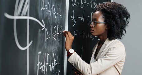 African American young woman teacher at school writing formulas and mathematics laws on blackboard....