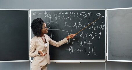 Young African American woman teacher in glasses standing at board in classroom and telling physics...
