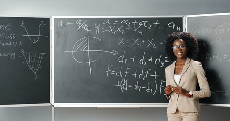 Rear of African American young female teacher of math or physics writing formulas on blackboard...