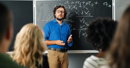 Caucasian man teacher at school at blackboard talking with pupils or students and asking question. Math class concept. Male lecturer in glasses explaining mathematics laws to kids. Educational concept