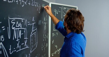 Caucasian male teacher in glasses and medical mask writing formulas and math laws in class. School concept. Educative mathematics lection. Close up of man lecturer turning to camera and looking.
