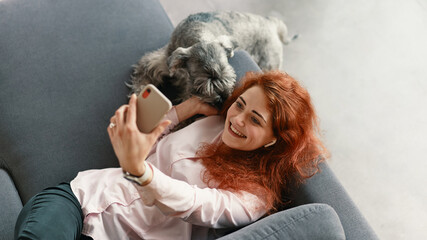 Top view of young happy woman relaxing on the sofa with her dog while making a video call