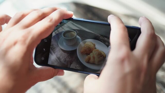 Closeup view 4k video of two female hands holding modern smartphone. Woman takes mobile photos of tasty breakfast cup of espresso coffee and two croissants on table to share in social media resaurces.