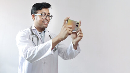 male indian doctor in white coat and stethoscope clicking photo with a mobile tripod and futuristic...